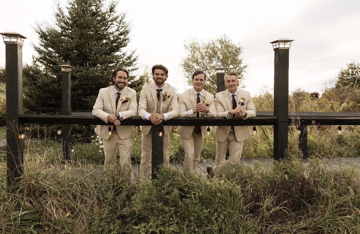 Groomsmen leaning along a wooden bridge railing in tan suits